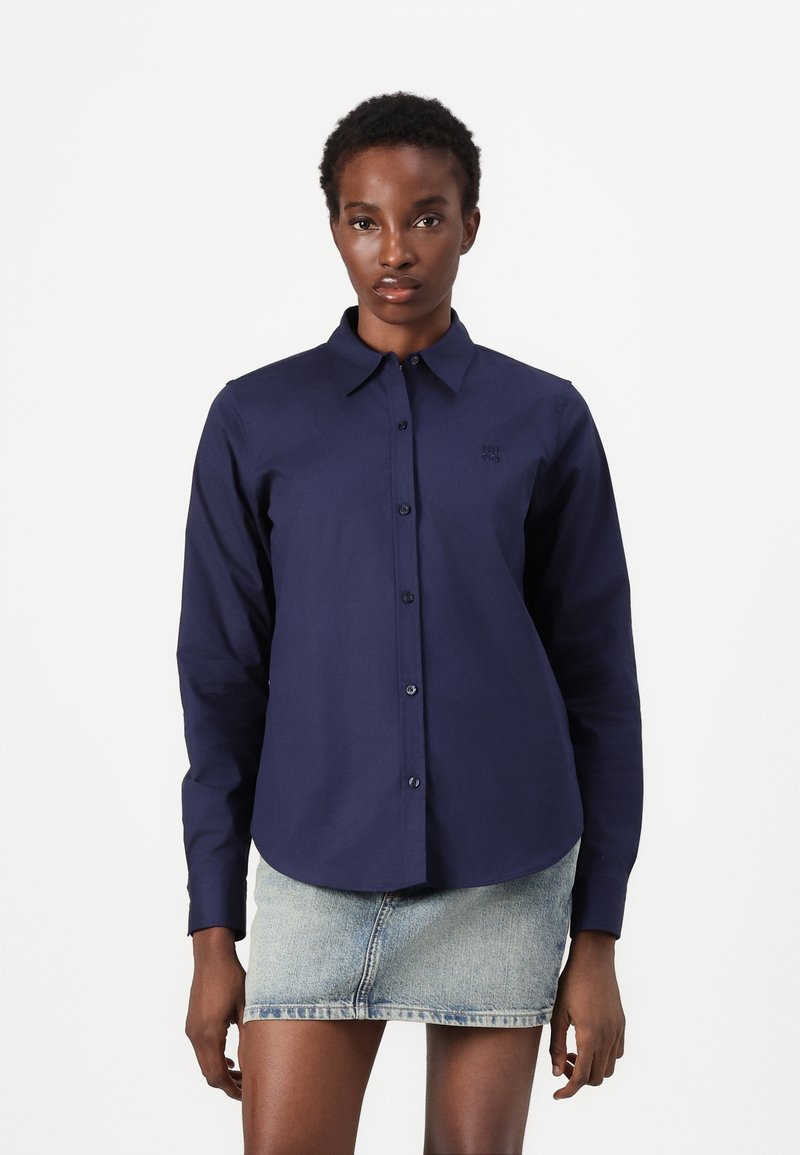 Camicia button-up a maniche lunghe di colore blu navy, realizzata in cotone, con colletto, dettaglio logo discreto e orlo curvo. Indossata con una gonna di jeans.
