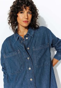 Chemise en denim bleu profond, avec un col, une fermeture à boutons, deux poches poitrine et des manches retroussées, offrant une coupe décontractée et un tissu texturé.