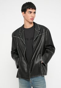 Noor mees kannab musta nahktagi ja musta särki, seljas pleekinud sinised teksapüksid, vaatab küljele, käed taskutes.