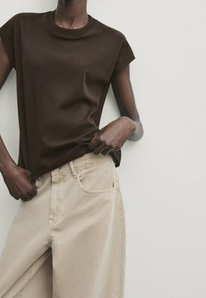 Person wearing a sleeveless dark brown top and loose beige pants, holding the hem of the top with one hand.