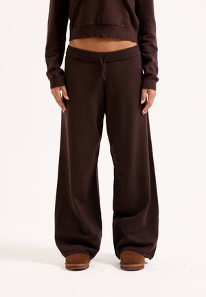 Maglione corto marrone con cappuccio e pantaloni della tuta a gamba larga abbinati, dotati di cintura con coulisse e una texture morbida. Le pantofole marroni completano il look.