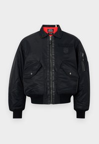 Veste bomber noire avec fermeture éclair à l'avant, deux poches à rabat, poche zippée sur la manche, poignets et ourlet côtelés, et doublure intérieure rouge.