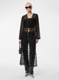 Soprabito in pizzo nero stile kimono con maniche lunghe, apertura frontale, motivo floreale, abbinato a un top nero e jeans grigi. Scarpe e borsa argentate.