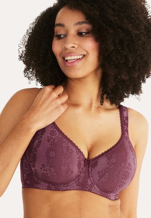 Lachende vrouw met krullend haar die een paarse bloemen kanten beha draagt, met haar rechterhand een bandje vasthoudend, kijkt iets naar beneden en opzij.