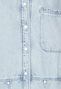 Camisa de mezclilla azul claro con botones en el frente, botones blancos, un bolsillo en el pecho y detalles de costura bien hechos. Textura suave con un diseño casual.