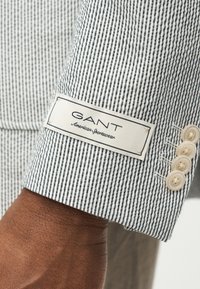 Close-up van een pols met een mouw van een zwart-wit gestreept GANT American Sportswear jasje met vier beige knopen.