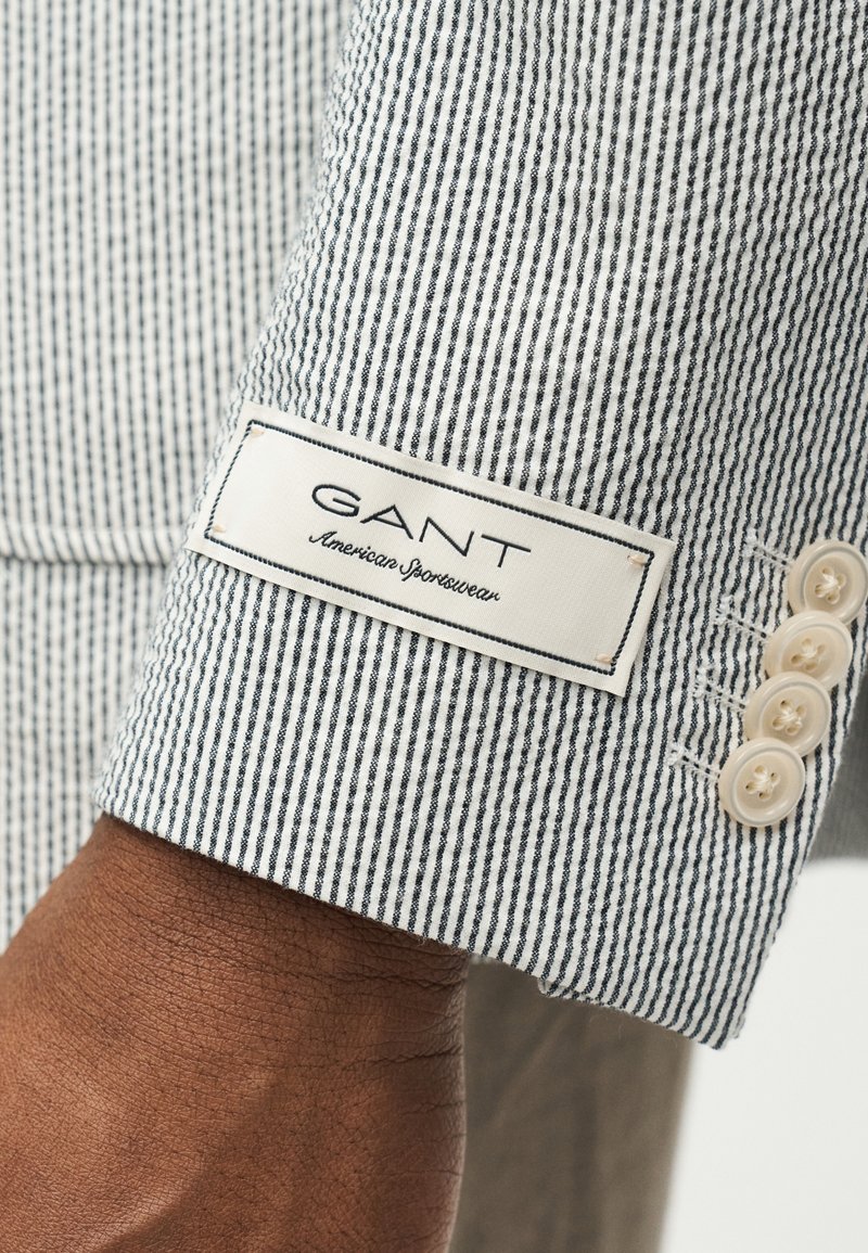 Primo piano del polso che indossa la manica di una giacca GANT American Sportswear a righe bianche e nere con quattro bottoni beige.