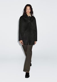 Czarny płaszcz z mieszanki wełny o strukturalnym, lekko A-liniowym fasonie, z jednym guzikem z przodu i bocznymi kieszeniami, w połączeniu z oliwkowymi spodniami.