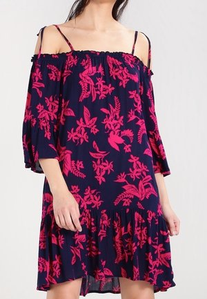 Kvinde iført en off-shoulder marineblå kjole med lyserødt blomsterprint og løstsiddende trekvartærmer.