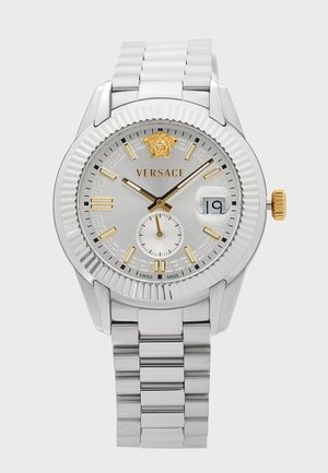 Montre-bracelet Versace argentée avec des accents dorés, cadran blanc, fenêtre date affichant 19 et bracelet à maillons métalliques.