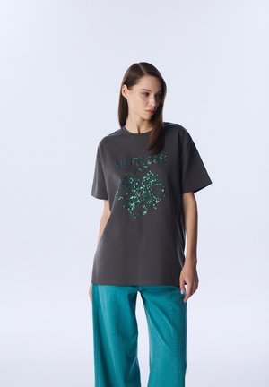 Grijze katoenen T-shirt met korte mouwen met een groene paillettenopdruk "LIMITLESS" op de voorkant, gecombineerd met turquoise wijde broek.