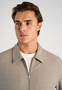 Jasnoszara kurtka na zamek z kołnierzem, wykonana z teksturowanej tkaniny. Nałożona na biały t-shirt. Gładkie wykończenie.