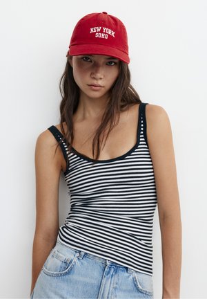 Junge Frau mit roter Mütze mit der Aufschrift "New York Soho", schwarz-weiß gestreiftem Tanktop und hellblauen Jeans vor einem weißen Hintergrund.