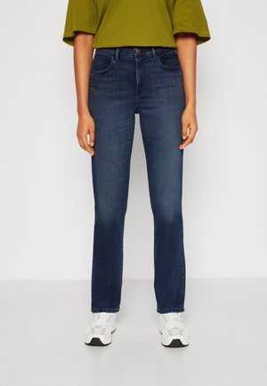 Osoba ubrana w ciemnoniebieskie jeansy o prostych nogawkach, zieloną koszulkę z krótkim rękawem i białe trampki, stojąca na jednolitym białym tle.