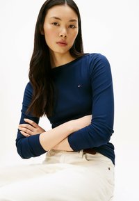 Marineblaues, geripptes Langarmoberteil mit Rundhalsausschnitt und kleinem Logo-Detail. Kombiniert mit hellen, strukturierten Hosen.