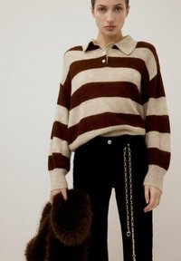 Suéter a rayas marrón y crema con cuello tipo polo, combinado con pantalones negros que tienen detalles de cadenas, sosteniendo un accesorio de piel oscura.