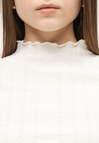 Κοντινό πλάνο του κάτω μέρους του προσώπου και του λαιμού ενός ατόμου που φοράει λευκή ριμπ υφασμάτινη μπλούζα με βολάν και ψηλό γιακά.