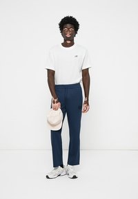 Tricou alb cu un logo mic, pantaloni albaștri cu o aplicație logo și o șapcă bej ținută în mână; pantofii sport completează ținuta.