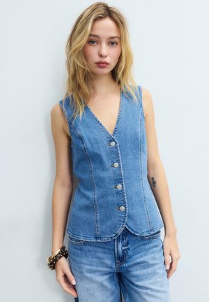 Młoda kobieta o blond włosach, ubrana w bezrękawnik z dżinsu zapinany na guziki oraz dopasowane jeansy, stojąca na tle białego, jednolitego tła.