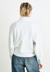 Lys grå hettegenser med høy krage, ribbede mansjetter og kant. Myk stoff med en avslappet passform, vist fra baksiden. Jeansene er mørkeblå, med et lapp.