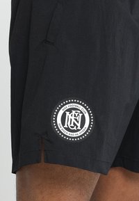 Sorte sorte shorts med et rundt hvidt Nike Football Club-logo og sætningen "Sem risco não há vitória" nær kanten på en persons lår.