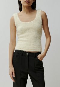 Ecru kleurige mouwloze top met een zachte, fluffy textuur, ronde halslijn en aansluitende silhouet, gecombineerd met zwarte broek met voorzakken.