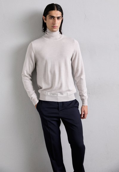Maglione a collo alto beige chiaro in una morbida texture, abbinato a pantaloni sartoriali blu scuro, indossato con le mani in tasca su uno sfondo neutro.