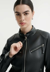 Černá bunda z umělé kůže s vysokým límcem, zipem vpředu a žebrovanými detaily na ramenou. Obsahuje dvě zipové kapsy. Hladký povrch.