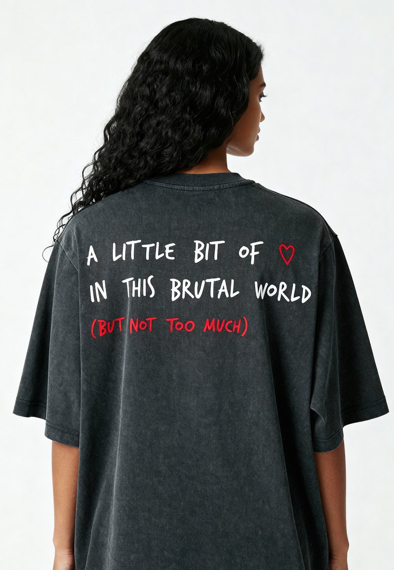Tricou negru din bumbac supradimensionat, cu un finisaj estompat. Text pe spate în alb și roșu: „Puțin ❤️ în această lume brutală (dar nu prea mult).”