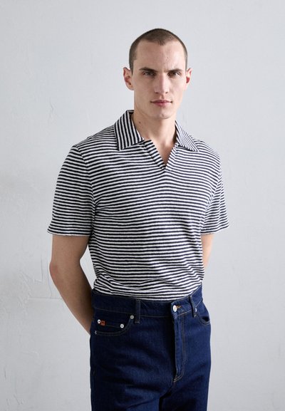 Polo de rayas de manga corta en azul marino y blanco, con un cuello ancho y un corte entallado. Lleva puesto unos jeans de denim oscuros, mostrando un estilo casual.