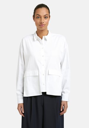 Woman with tied-back hair wearing a white button-up shirt with front pockets and black loose trousers, standing against a plain background.