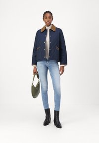 Granatowa pikowana kurtka z brązowym kołnierzem z kordurowej tkaniny, pasującymi akcentami na kieszeniach, jasny sweter, niebieskie dżinsy oraz czarne botki na obcasie, trzymająca zieloną torbę.