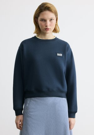 Jeune femme aux cheveux blonds courts portant un sweat-shirt bleu marine et une jupe à rayures bleues, debout devant un fond blanc uni.