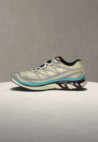 Ανοιχτό μπεζ και γκρι αθλητικό παπούτσι με λευκά κορδόνια, τιρκουάζ και σκούρα μωβ ενδιάμεση σόλα και λογότυπο "Salomon" στο πλάι.