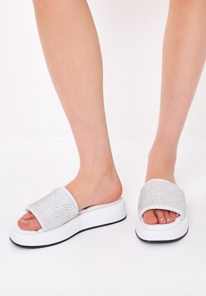 Pieds portant des sandales blanches à plateforme avec des brides couvertes de strass sur un fond blanc uni.