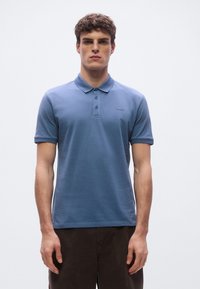 Zils kokvilnas polo krekls ar apkakli, trim pogām un zīmola "HUGO" izšuvumu uz krūtīm. Modelis stāv taisni.