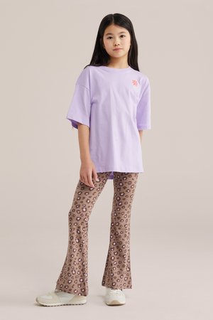 Niña joven vestida con una camiseta lavanda holgada con un logo naranja y pantalones acampanados de estampado de leopardo marrón, de pie frente a un fondo liso.