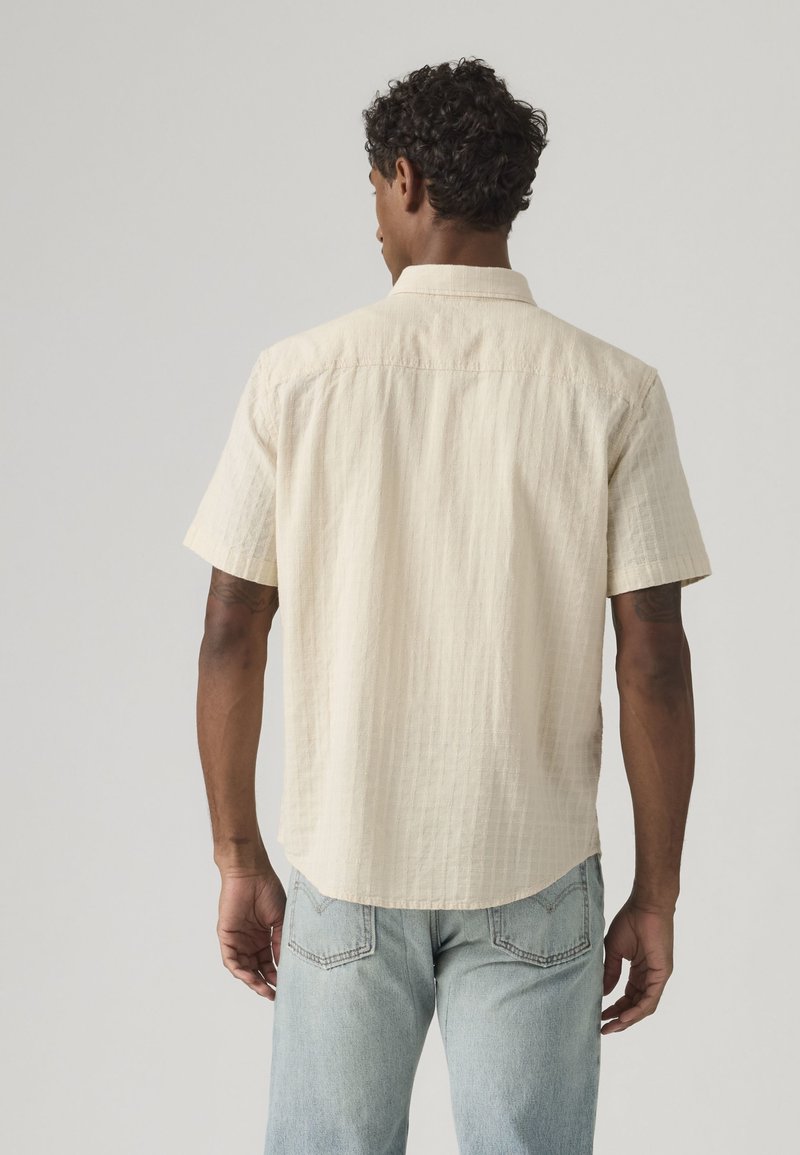 Homme aux cheveux courts et bouclés, portant une chemise texturée à manches courtes crème et un jean bleu clair, vu de dos sur un fond uni.