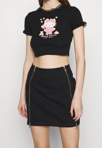 Mladá žena v černém zkráceném tričku Hello Kitty s krajkovým lemem a černé minisukni se dvěma předními zipy.