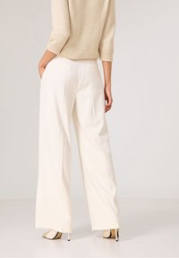 Pantalons larges blancs en tissu léger et lisse, avec des poches latérales et une coupe soignée à la taille, associés à un pull beige texturé.