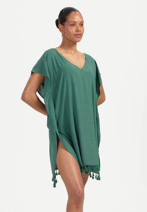 Femme debout, mains dans le dos, portant une robe verte ample à col en V avec des fentes latérales et des franges à l'ourlet, sur un fond blanc.