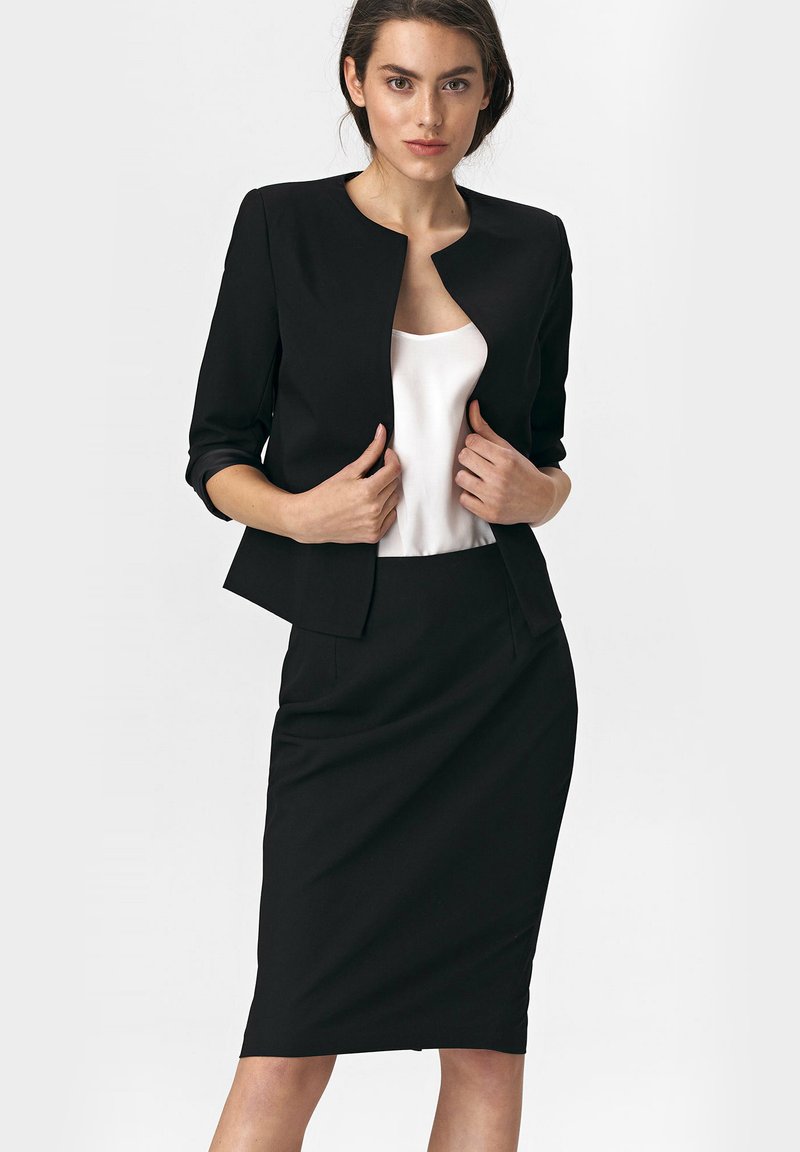 Kobieta w czarnym żakiecie i ołówkowej spódnicy z białą bluzką, trzymająca brzegi żakietu, na tle białego, gładkiego tła.