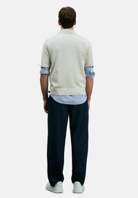Hellbeige Kurzarm-Polo aus Strick, drüber getragen über einem blau-weißen gestreiften Hemd. Dunkelblaue weite Hose und weiße Sneaker runden das Outfit ab.