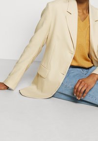 Personne assise par terre portant un blazer crème sur un pull moutarde et un jean bleu clair, le bras posé au sol.