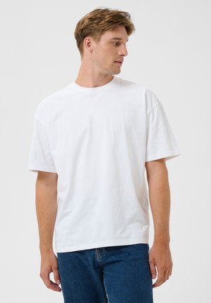 Joven con camiseta blanca lisa de manga corta y vaqueros azules mirando hacia su derecha sobre un fondo gris claro.