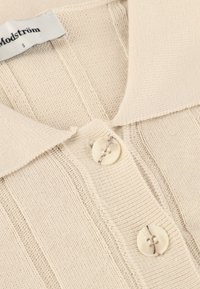 Lys beige strikket top med kravedesign og tre knapper, der fremhæver lodrette ribber og en blød tekstur. Brandlabel synlig.