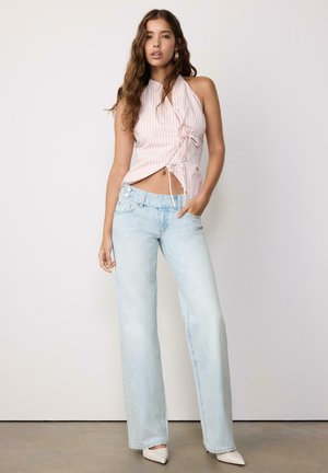 Kobieta z długimi włosami, ubrana w różowy top w paski bez rękawów z wiązaniami po bokach, jasnoniebieskie szerokie spodnie jeansowe i białe buty na czubku, stojąca.