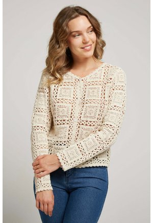 Femme souriante aux cheveux ondulés portant un haut crème en crochet à manches longues et un jean bleu, debout devant un fond clair uni.