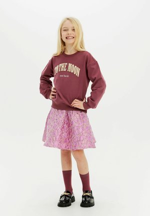 Mörkröd sweatshirt med texten "TO THE MOON and back" i kombination med en glänsande rosa utsvängd kjol, burgundy-strumpor och svarta skor med kedjedetalj.