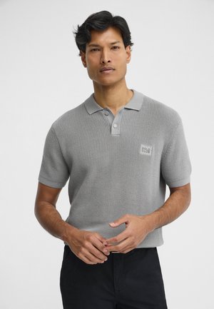 Mężczyzna w szarej, krótkorękawowej dzianinowej koszulce polo z guzikami i czarnych spodniach, stoi na tle jednolitego białego tła.